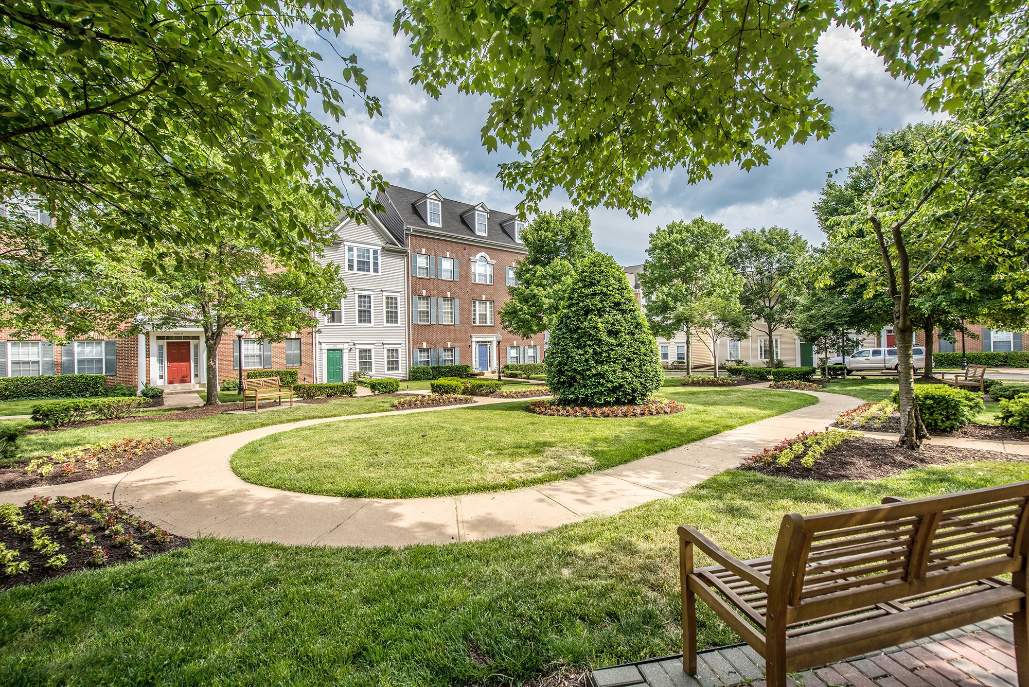 Beautifully landscaped apartment community courtyard featuring circular walkway, trimmed hedges, and wooden benches