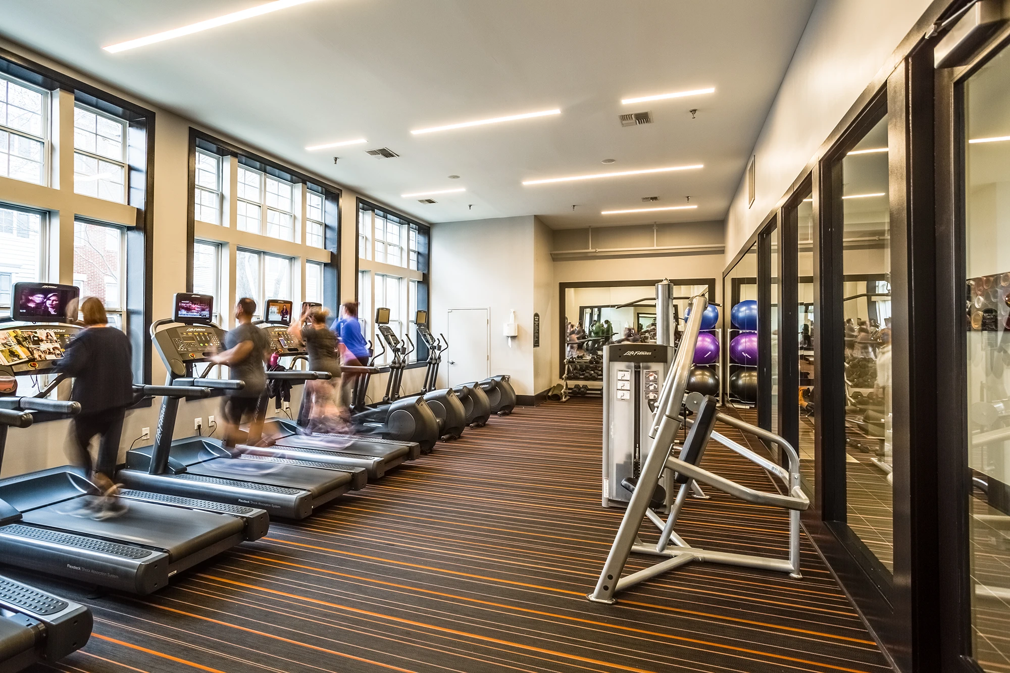 Bright apartment gym room with treadmills, elliptical machines, large mirrors, and striped flooring design
